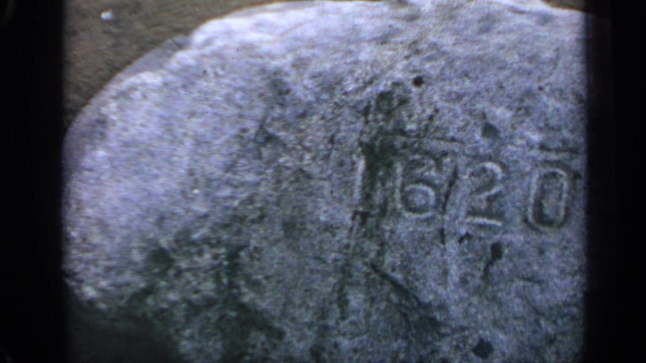 1969 Massachusetts home-movie frame with a close handheld view of a carved 6/20 marking on rough stone.