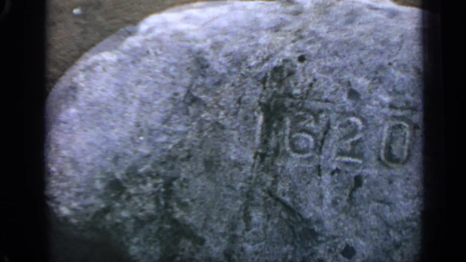 1969 Massachusetts home-movie frame with a close handheld view of a carved 6/20 marking on rough stone.
