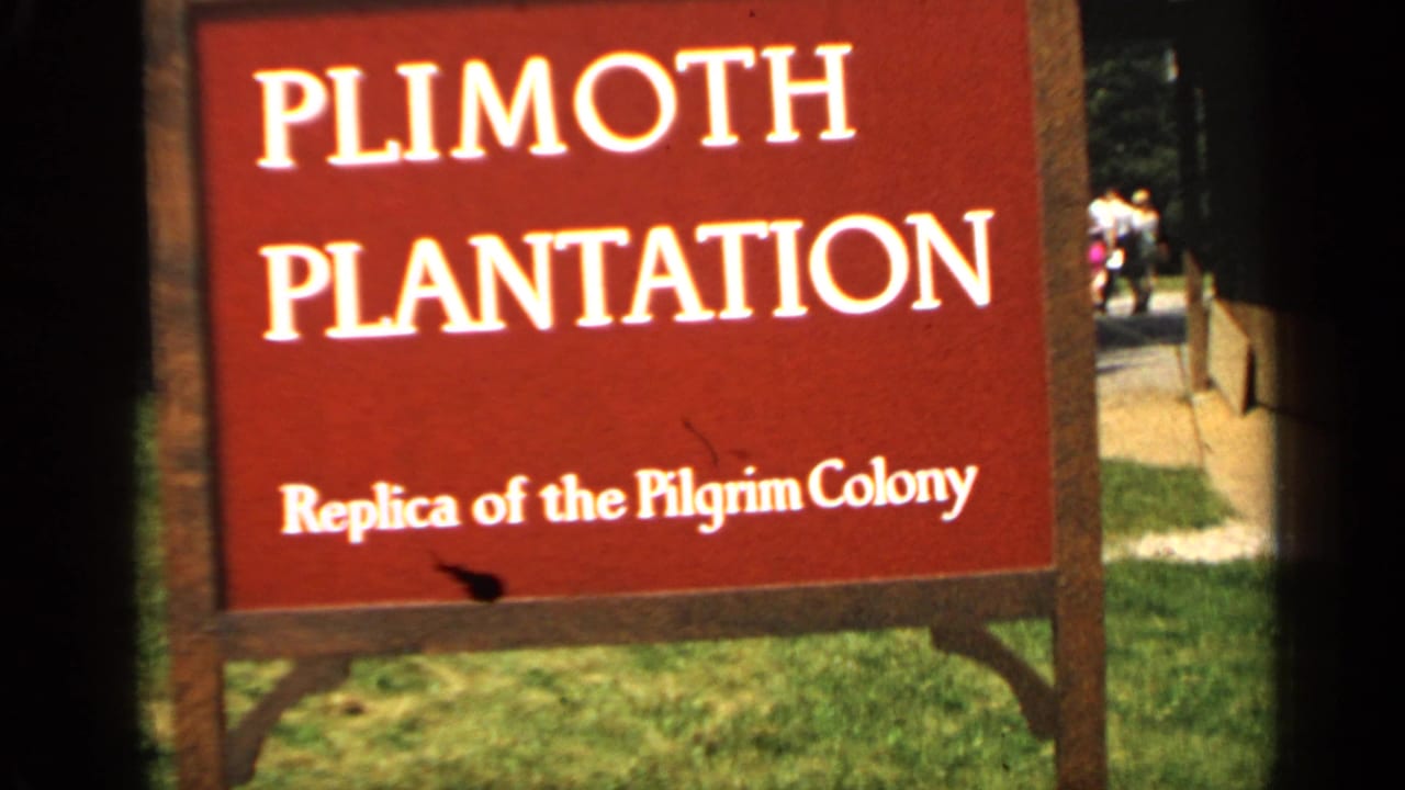 Red Plimoth Plantation sign for a replica of the Pilgrim Colony, with grass and distant visitors in Massachusetts.