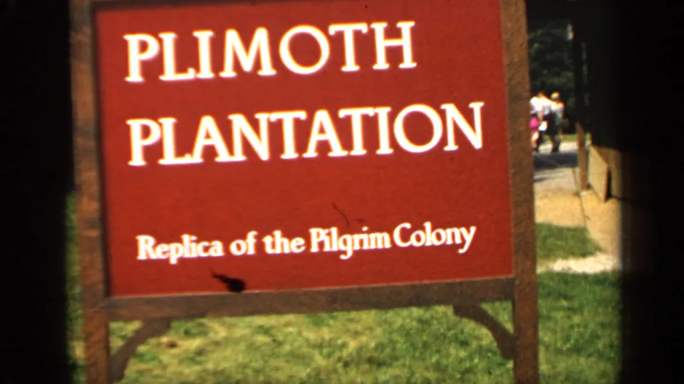 Red Plimoth Plantation sign for a replica of the Pilgrim Colony, with grass and distant visitors in Massachusetts.
