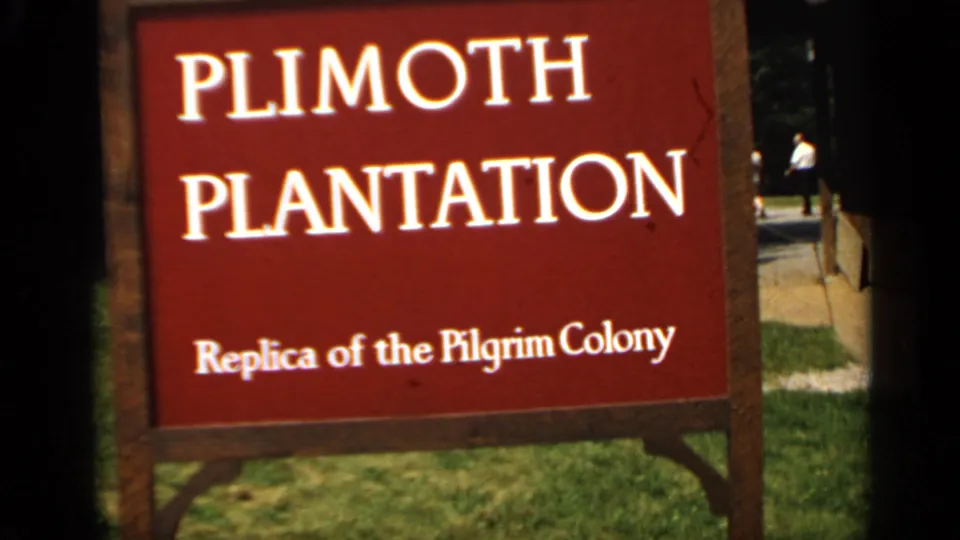 Close visitor-shot view of the Plimoth Plantation sign, with a path and distant person beyond the wooden frame.