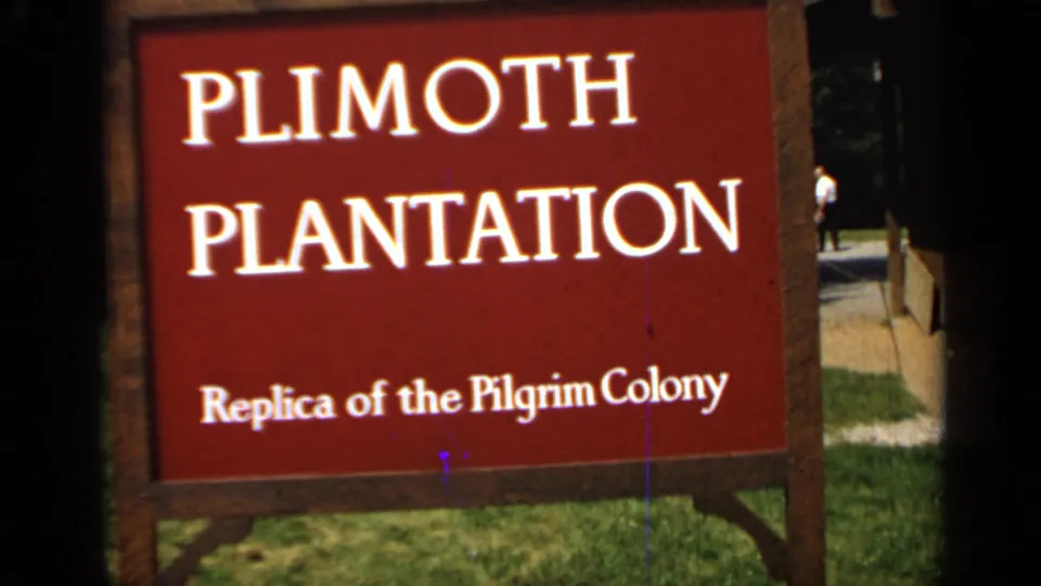 Handheld 8mm view of the red Plimoth Plantation sign, with grass, path, and distant visitors at the edge.