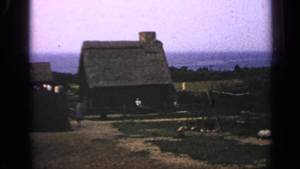 Wide 1969 Massachusetts home-movie view of a thatched-roof cottage, fence line, open ground, and small distant figures outdoors.