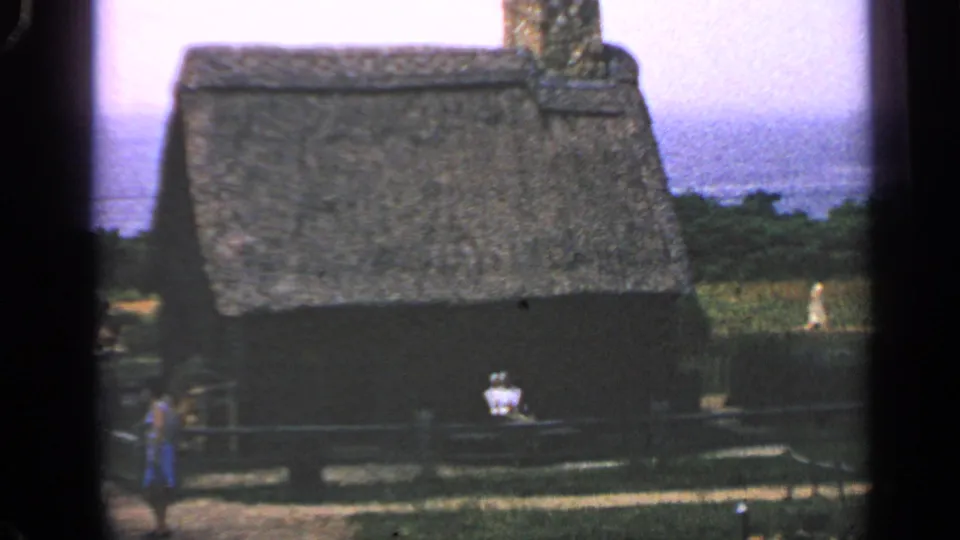 1969 Massachusetts frame showing a thatched cottage with a stone chimney, fence, and distant people near the building.