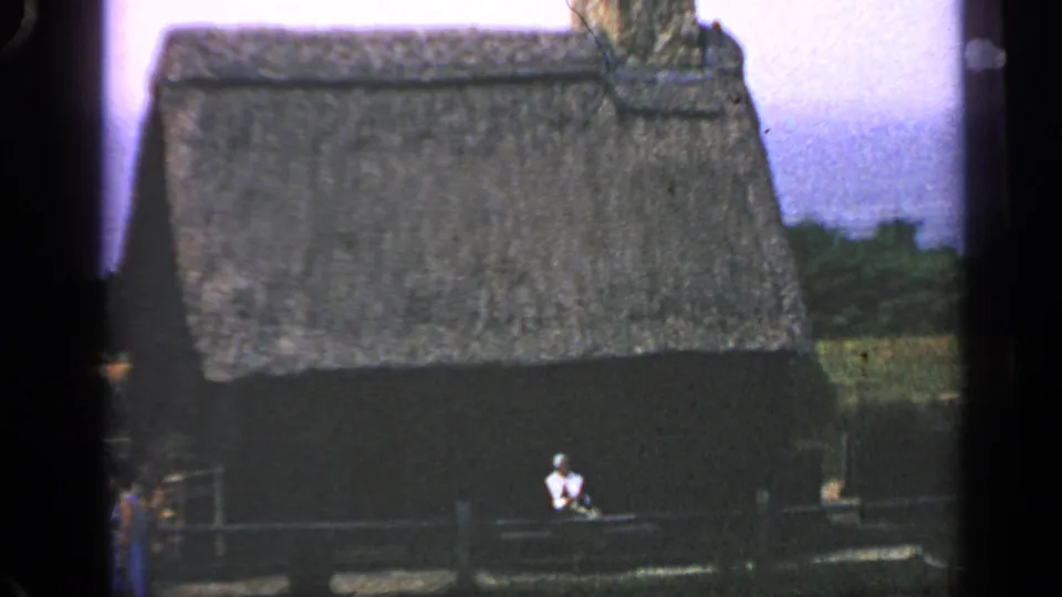 1969 Massachusetts 8mm frame showing a rustic thatched-roof cabin, fence, greenery, and a seated person in a white shirt.