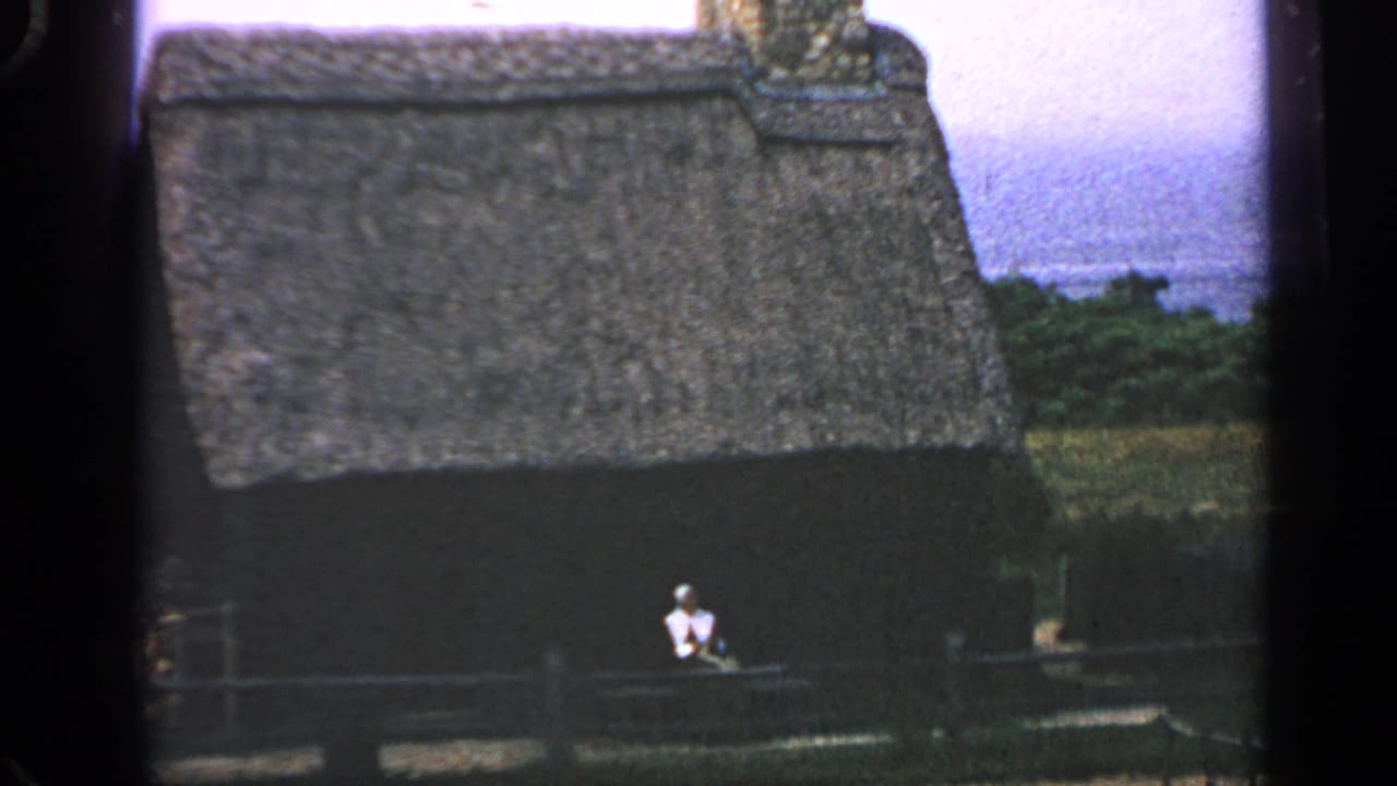 1969 Massachusetts home-movie frame with a thatched-roof cabin, dark wall, wooden fence, and seated figure in front.