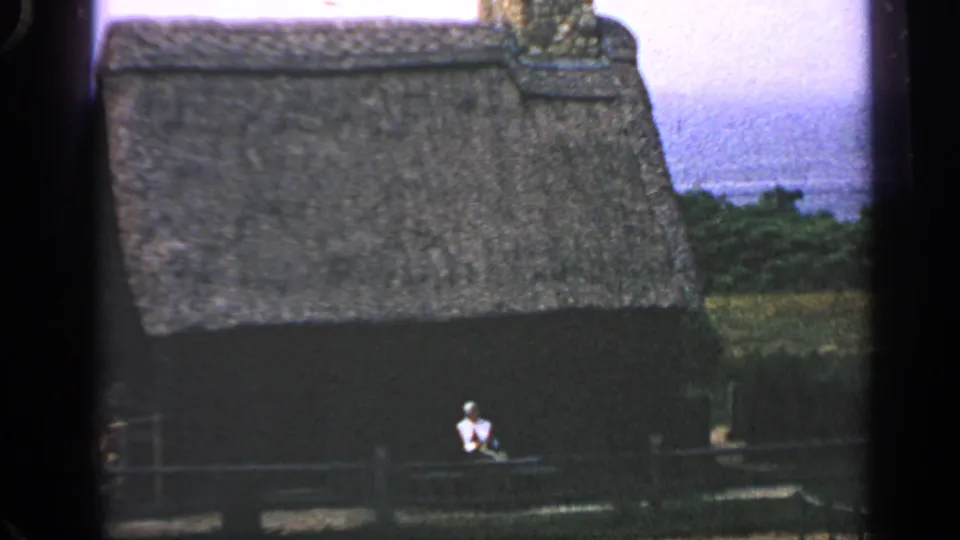 1969 Massachusetts home-movie frame with a thatched-roof cabin, dark wall, wooden fence, and seated figure in front.