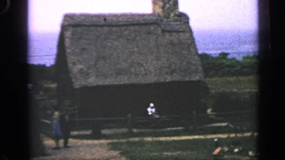 Wider 1969 Massachusetts 8mm view showing the thatched cabin, fence, seated person, and standing visitor at the left edge.