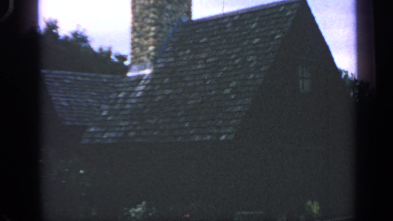1969 Massachusetts home-movie frame with a steep shingled roof, stone chimney, small window, and trees.