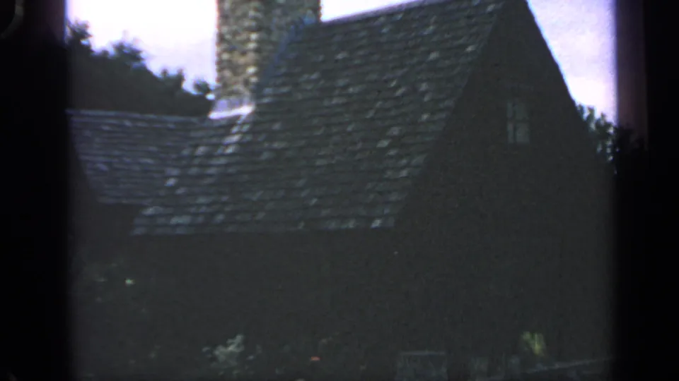 1969 Massachusetts 8mm frame of a rustic roof, chimney, small window, trees, and shadowed foreground.