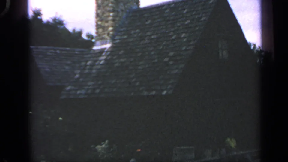 1969 Massachusetts frame showing a dark rustic house roof, stone chimney, small window, and wooded edge.