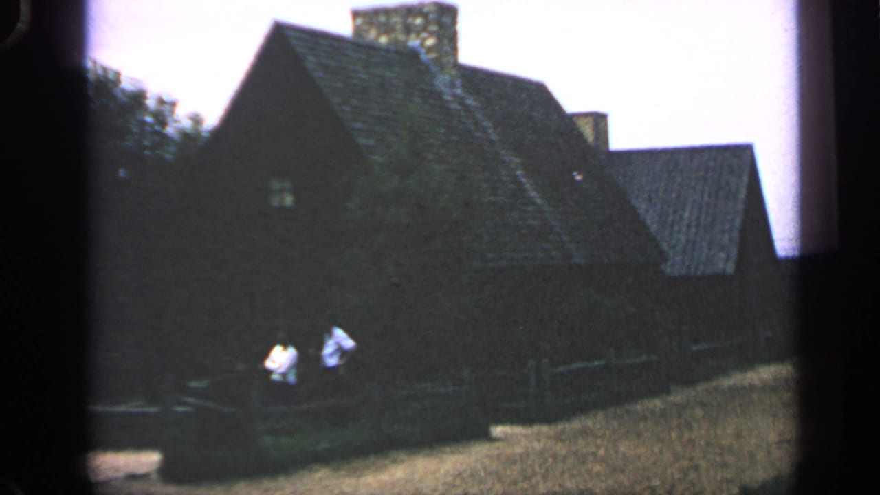 Massachusetts 1969 home-movie frame with two men in light shirts near a wooden fence before a dark shingled house.