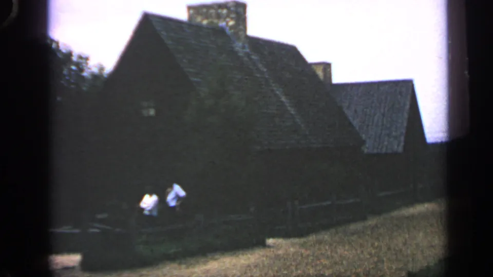 Massachusetts 1969 frame showing two men beside a fence, with a steep-roofed house and stone chimney behind them.