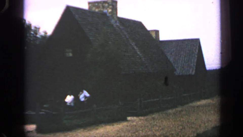 Massachusetts 1969 8mm frame with two men near a fence and a dark, steep-roofed house in the background.