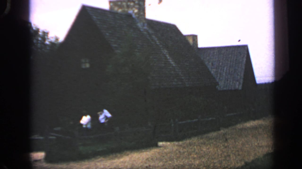 Dark wooden gabled building with stone chimney, fence, and two people in light clothing nearby.