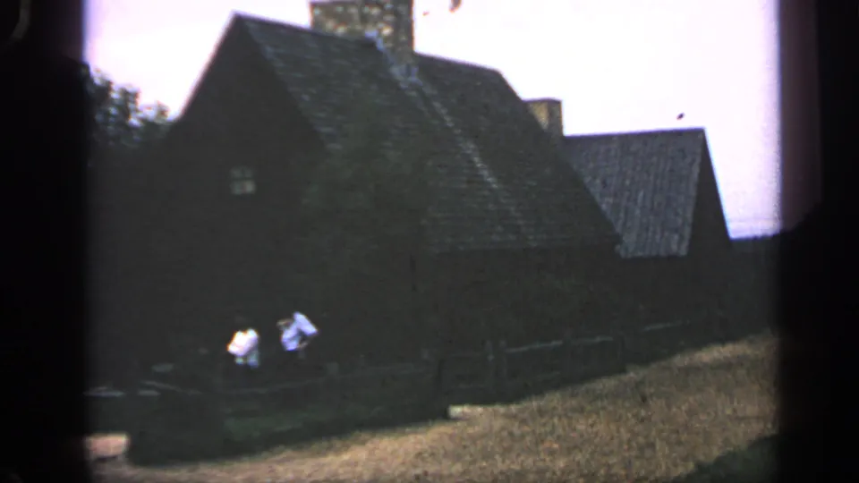 1969 Massachusetts 8mm frame showing a dark wooden historic-looking building, steep roof, stone chimney, fence, and two visitors in the foreground.