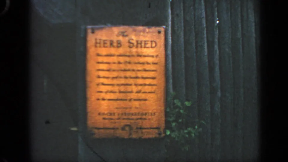 Close-up frame of the Herb Shed plaque on dark wooden siding, with readable heading and smaller interpretive text partly softened by 8mm focus.