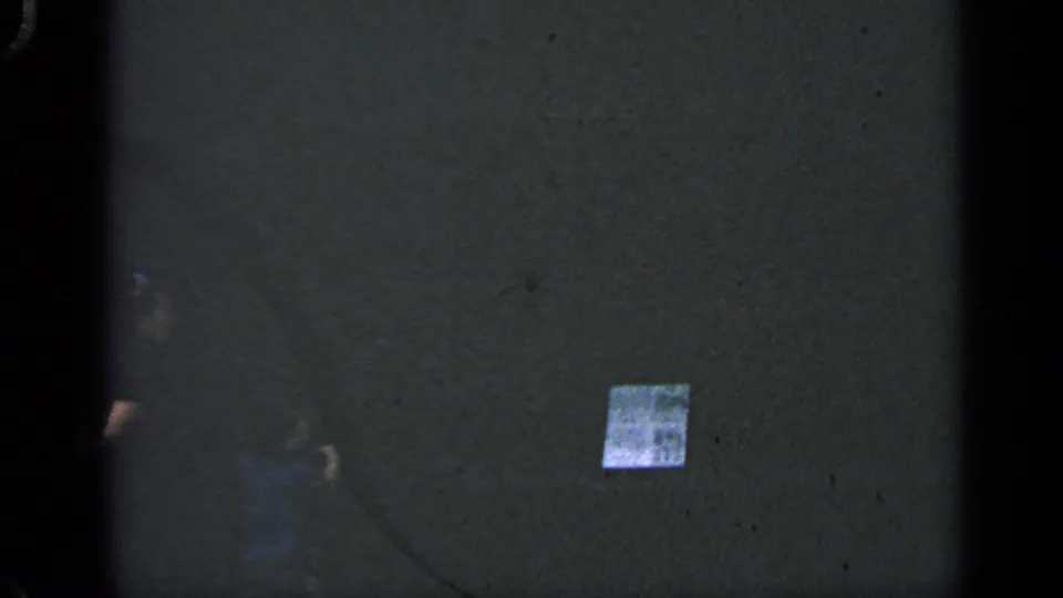 Final 8mm frame shows a mostly dark textured surface with a small reflective square, possibly a label or sticker, near the lower right.