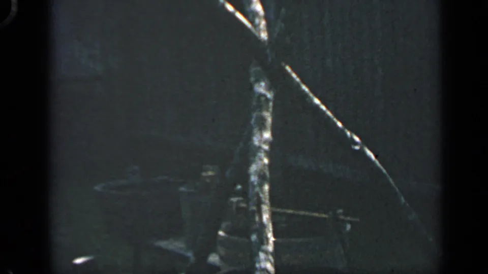 1969 Massachusetts 8mm frame with a tripod support, wooden vessels, and shaded exhibit-like surroundings.
