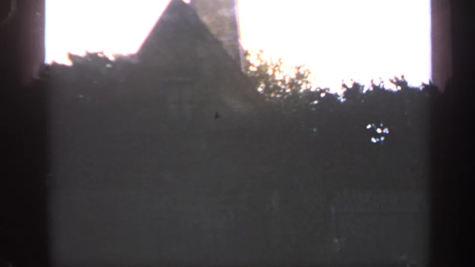 1969 Massachusetts 8mm frame with a steep stone roof or tower shape behind trees, partly obscured by dark gate shadow.