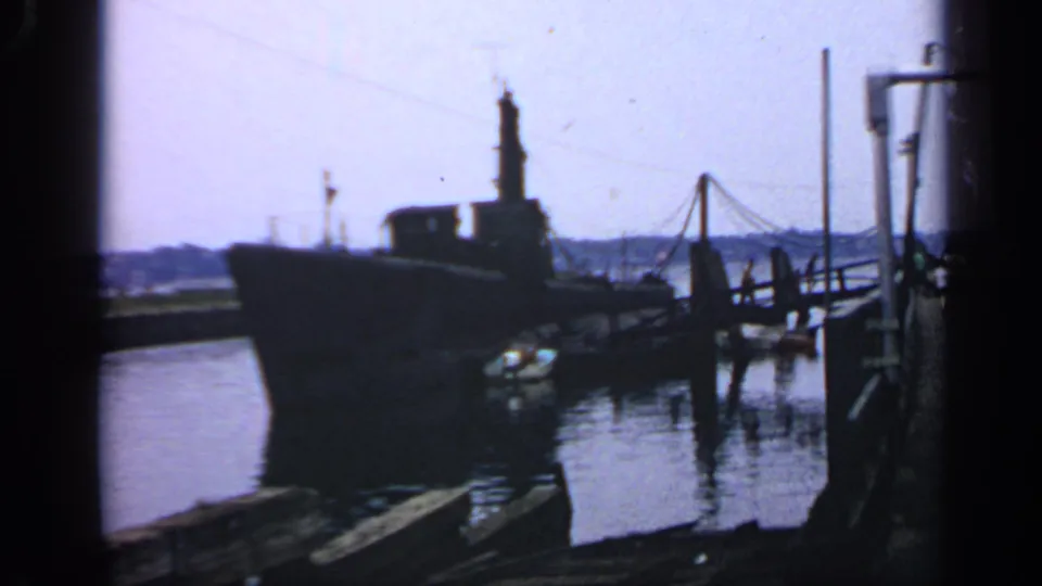 1969 Massachusetts 8mm frame showing a large submarine docked near a wooden pier with harbor water and distant shoreline.