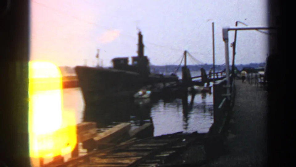 1969 8mm harbor frame with a docked submarine, wooden pier, calm water, and a strong yellow film-edge flare.