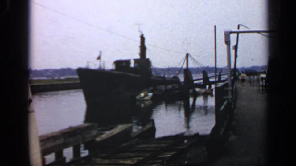Soft 8mm frame of a Massachusetts docked submarine, with wooden pier boards, harbor reflections, and small distant people on the dock.