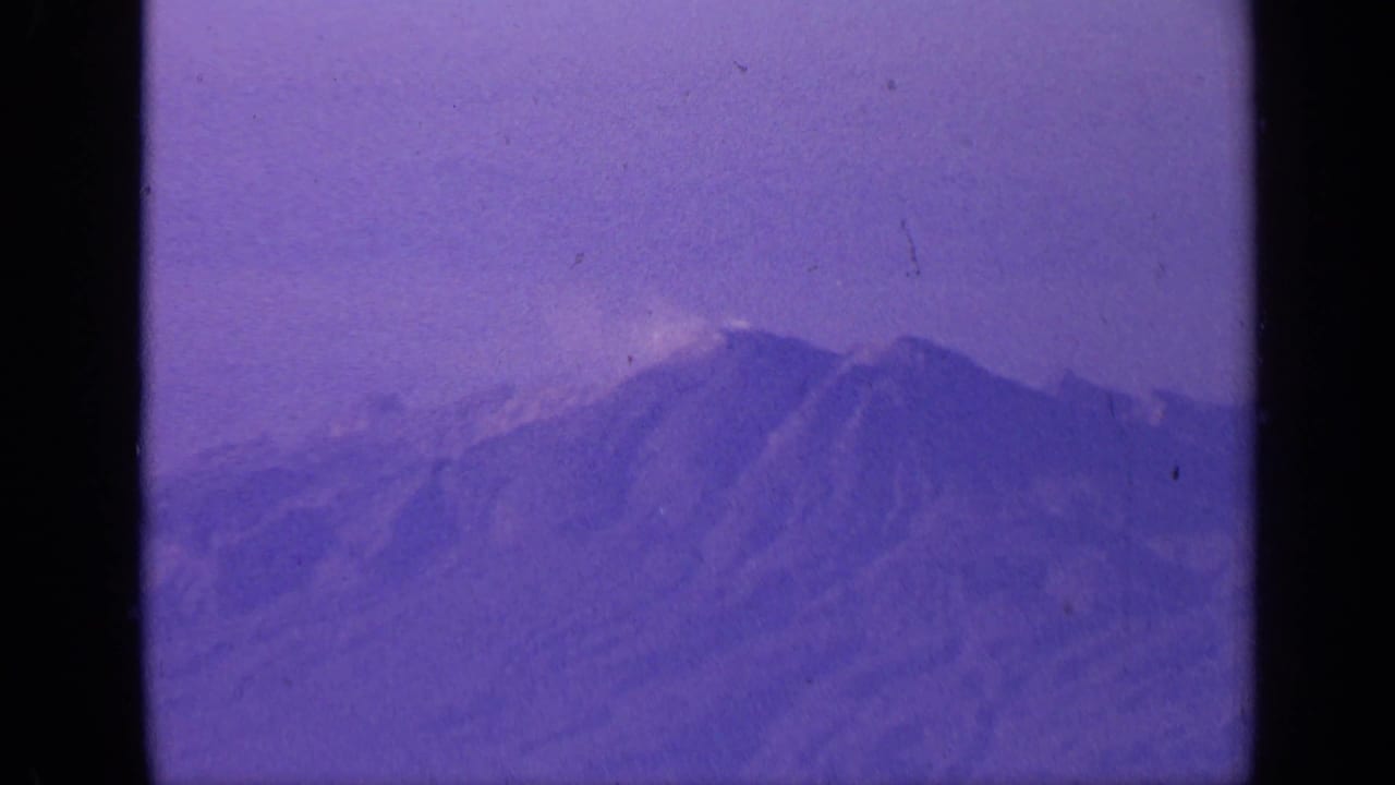 1970 India 8mm frame showing hazy snow-capped mountain peaks from a passenger aircraft, with purple color shift and film grain.