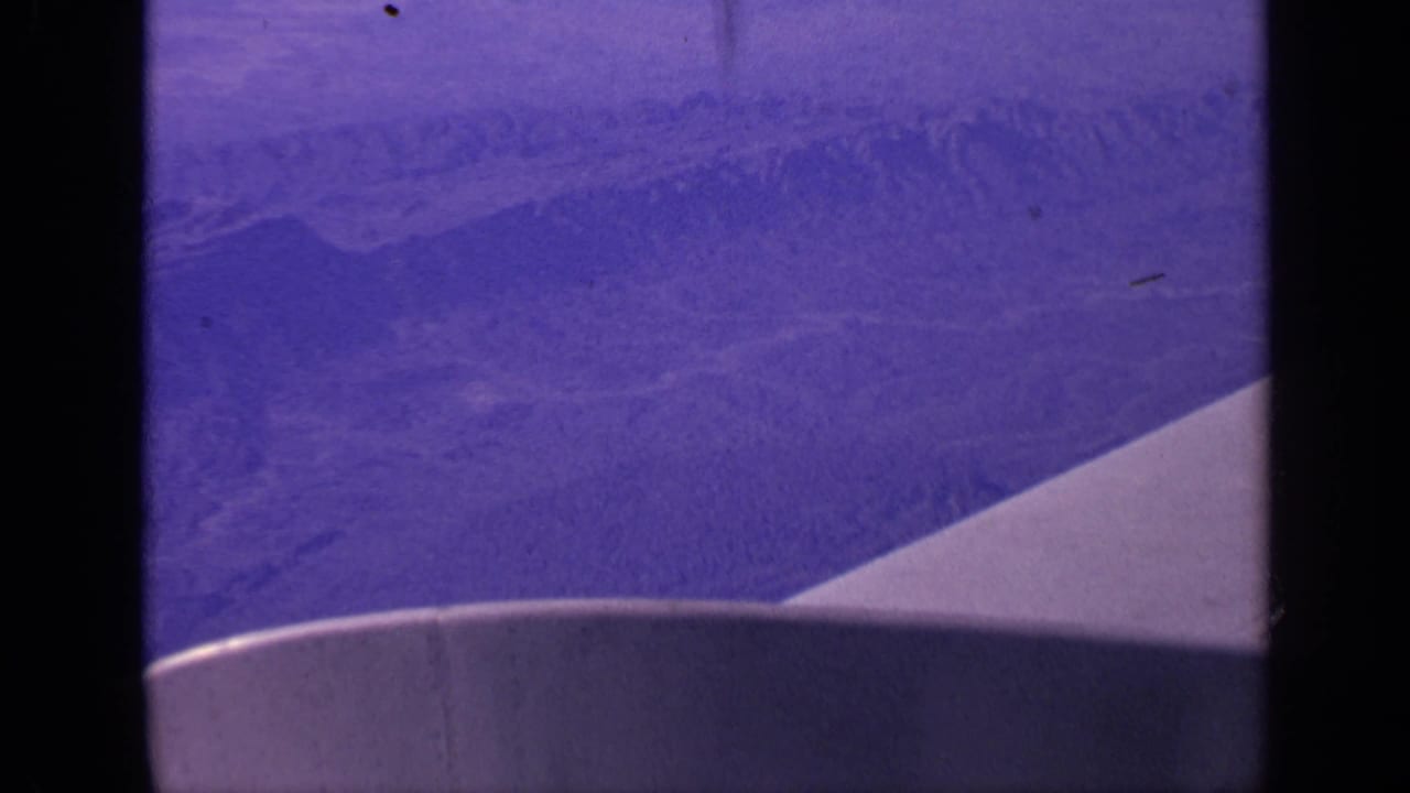 Airplane wing edge over hazy mountain ridges seen through a dark window frame.