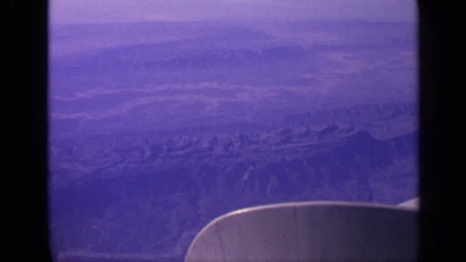 1970 India passenger-window frame showing a wing tip at lower right and ridged terrain softened by purple film fading.