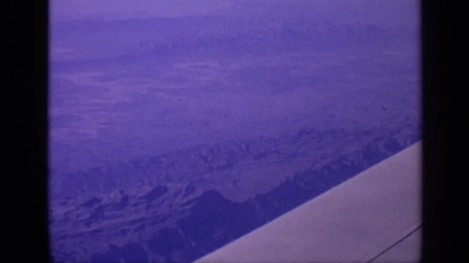 1970 India 8mm aerial view of hazy mountain ridges through an airplane window, with the aircraft wing crossing the foreground.