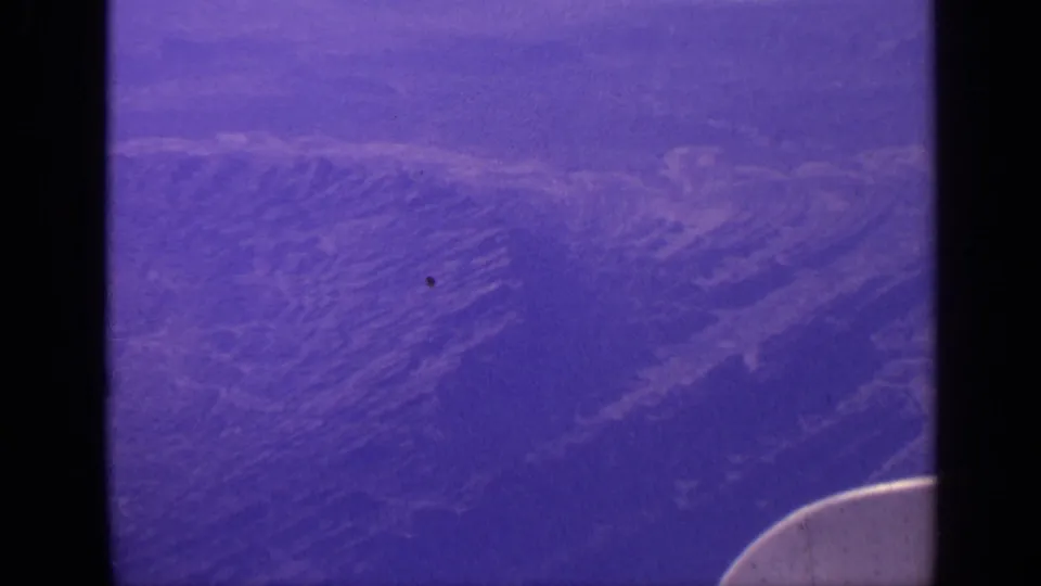 1970 India travel footage shows mountain ridges below a passenger airplane, with dark window framing and a blue-purple film cast.