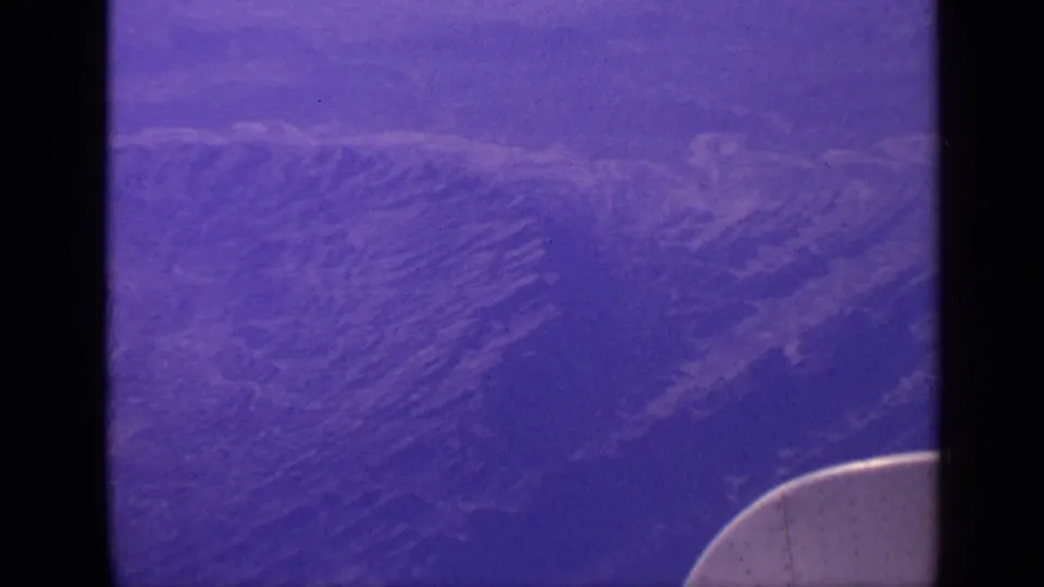 1970 India 8mm frame of mountain ridges seen from a passenger airplane, with window shadows and a curved aircraft edge visible.