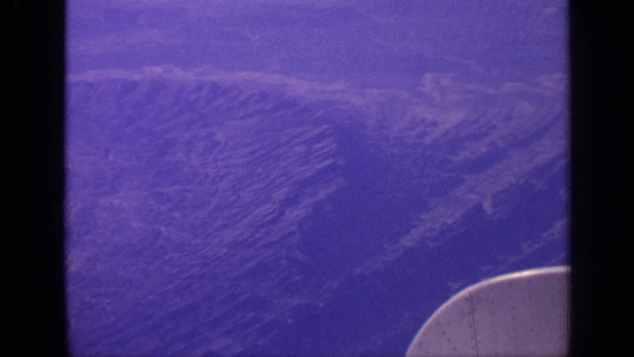 Purple-tinted aerial view of rugged mountain ridges through an airplane window.