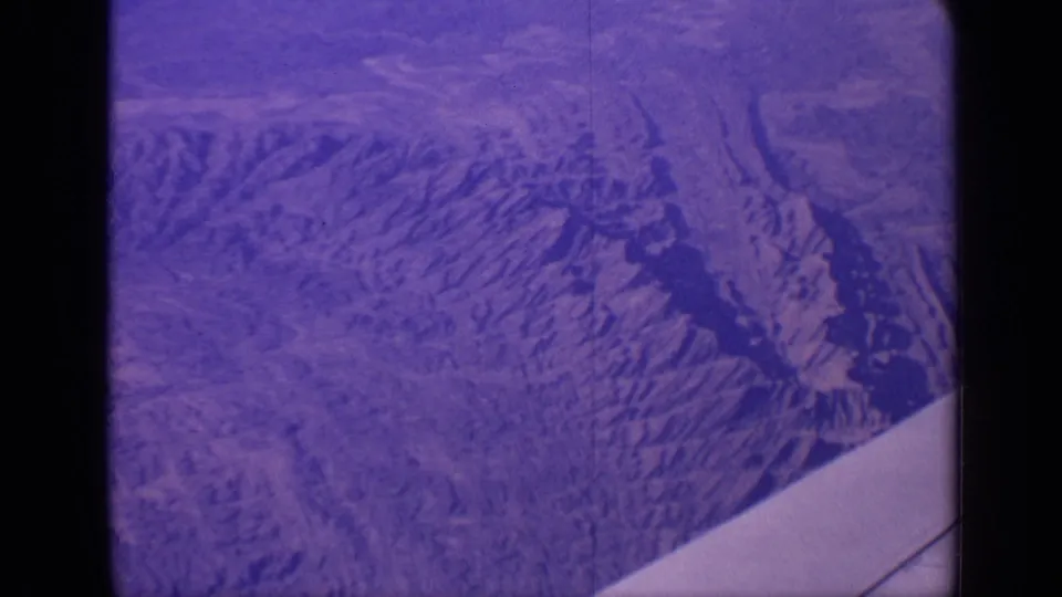 A purple-tinted 1970 India 8mm aerial frame shows ridged mountain terrain and valley shadows from a passenger-plane viewpoint.
