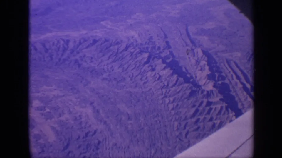 1970 India home-movie frame from an airplane window, showing rugged mountain ridges below and the aircraft wing along the lower right.
