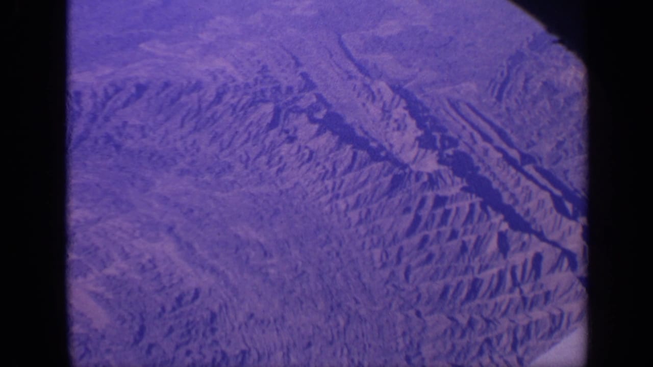 Purple-tinted aerial view of ridged mountains and dry valleys from an aircraft.