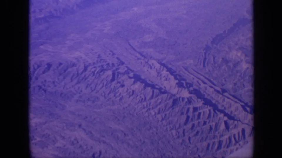 Faded 8mm aerial frame over India with ridged mountains, open arid ground, and visible film-frame borders.