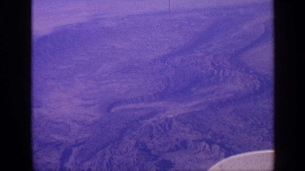 Purple-tinted aerial view of rugged mountain terrain with a curved aircraft wing edge at lower right.