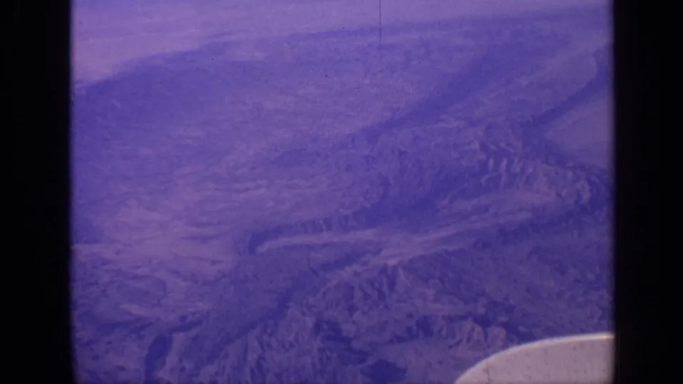 1970 India home-movie frame showing an aerial view over rugged terrain, cloud haze, purple color shift, and a partial aircraft wing.