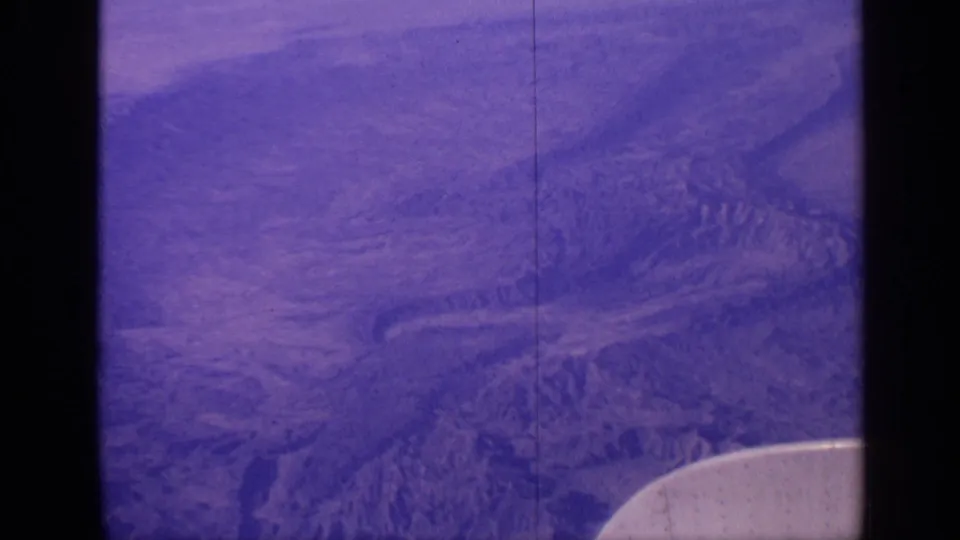 1970 India aerial home-movie still with purple-shifted mountain terrain, cloud haze, film scratches, and partial aircraft wing.