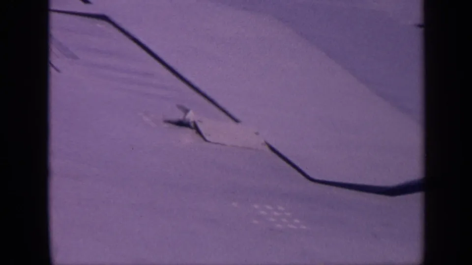 Airplane wing in flight, likely filmed from a passenger seat, with purple-blue tint and visible 8mm frame edges.