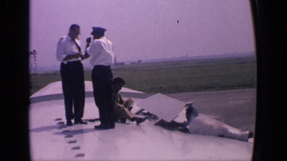 In 1970s India, two uniformed airline crew members appear to talk on a parked aircraft wing as a worker crouches beside parts or tools.