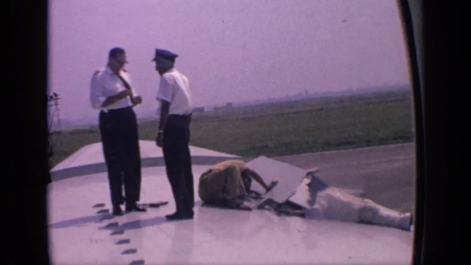 A uniformed crew pair stands on a parked aircraft wing at an Indian airfield, with maintenance items and a worker visible near the wing surface.