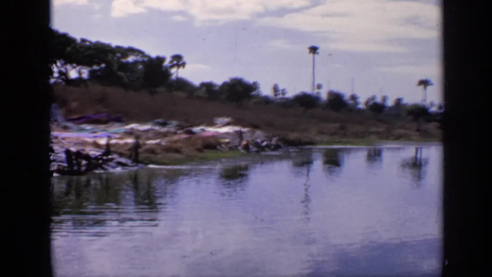 1970 India home-movie frame with reflective lake water, laundry spread near the shore, distant people, and palm trees on the horizon.