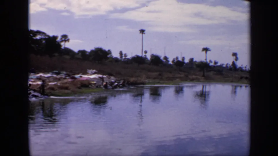 1970 India 8mm frame showing a lakeside bank with laundry, small figures near the water, and palm trees reflected in the lake.