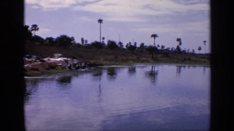 1970 India home-movie frame looking across open lake water to a low bank with laundry, distant people, grasses, and palms.