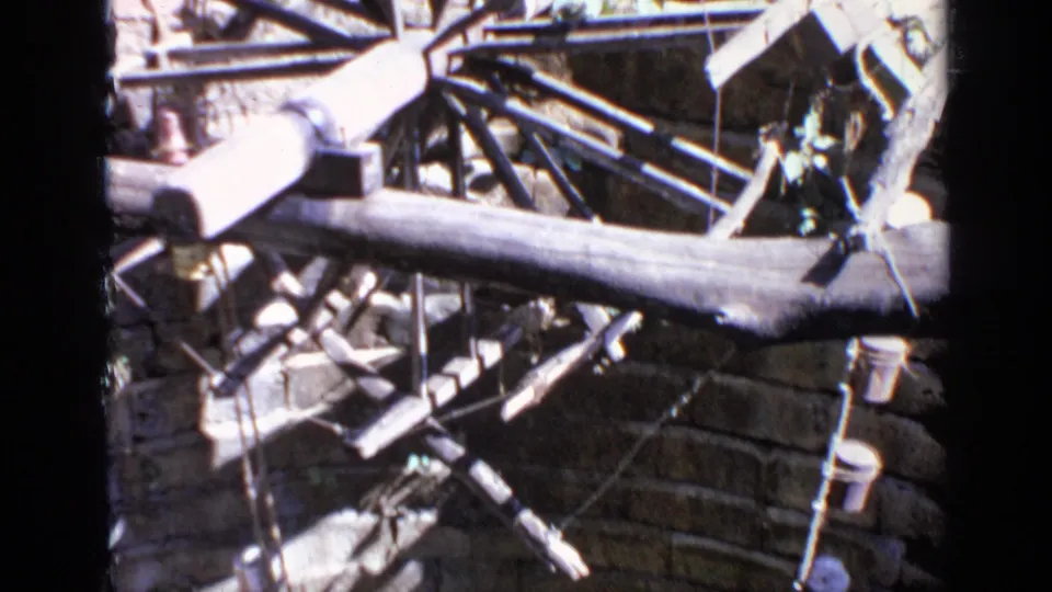 A rural 1970 India frame shows a spoked wooden wheel, beams, rods, buckets, and wall texture in harsh daylight.