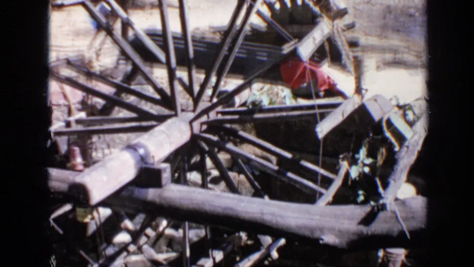 1970 India home-movie frame with a dark spoked wheel, pale wooden beam, rods, and scattered materials beside a wall.
