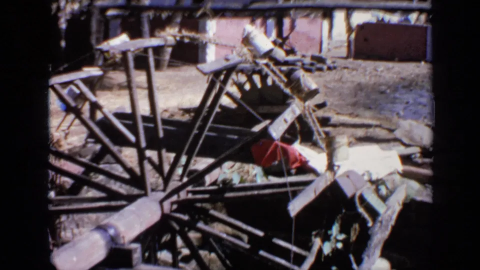 Wider 1970 India frame showing the wooden wheel assembly, beam, rough wall, hanging pieces, and ground debris.